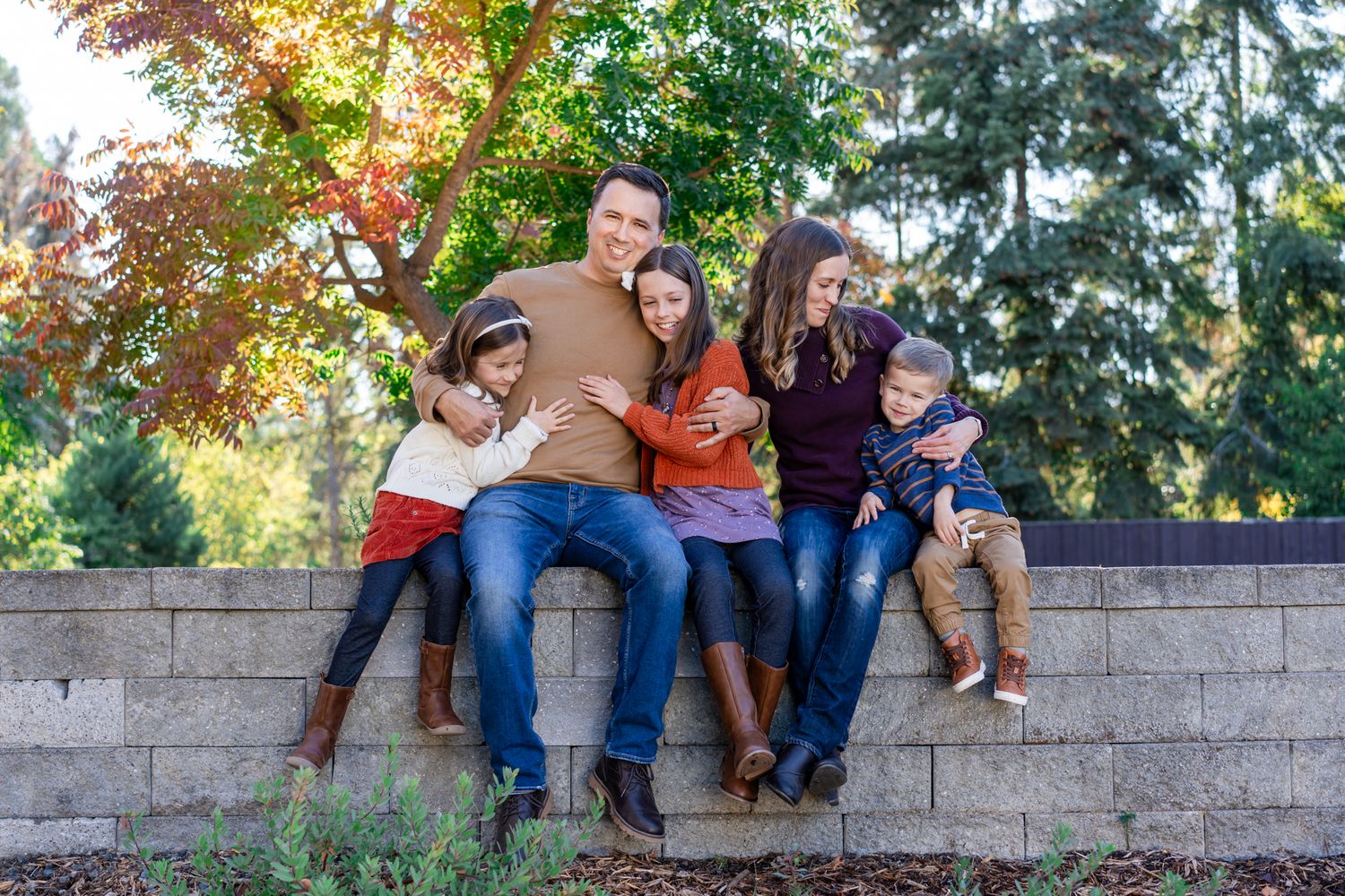 family of 5 sits on a retaining wall with fall-colored trees behind them for a family portrait in Jacksonville Oregon