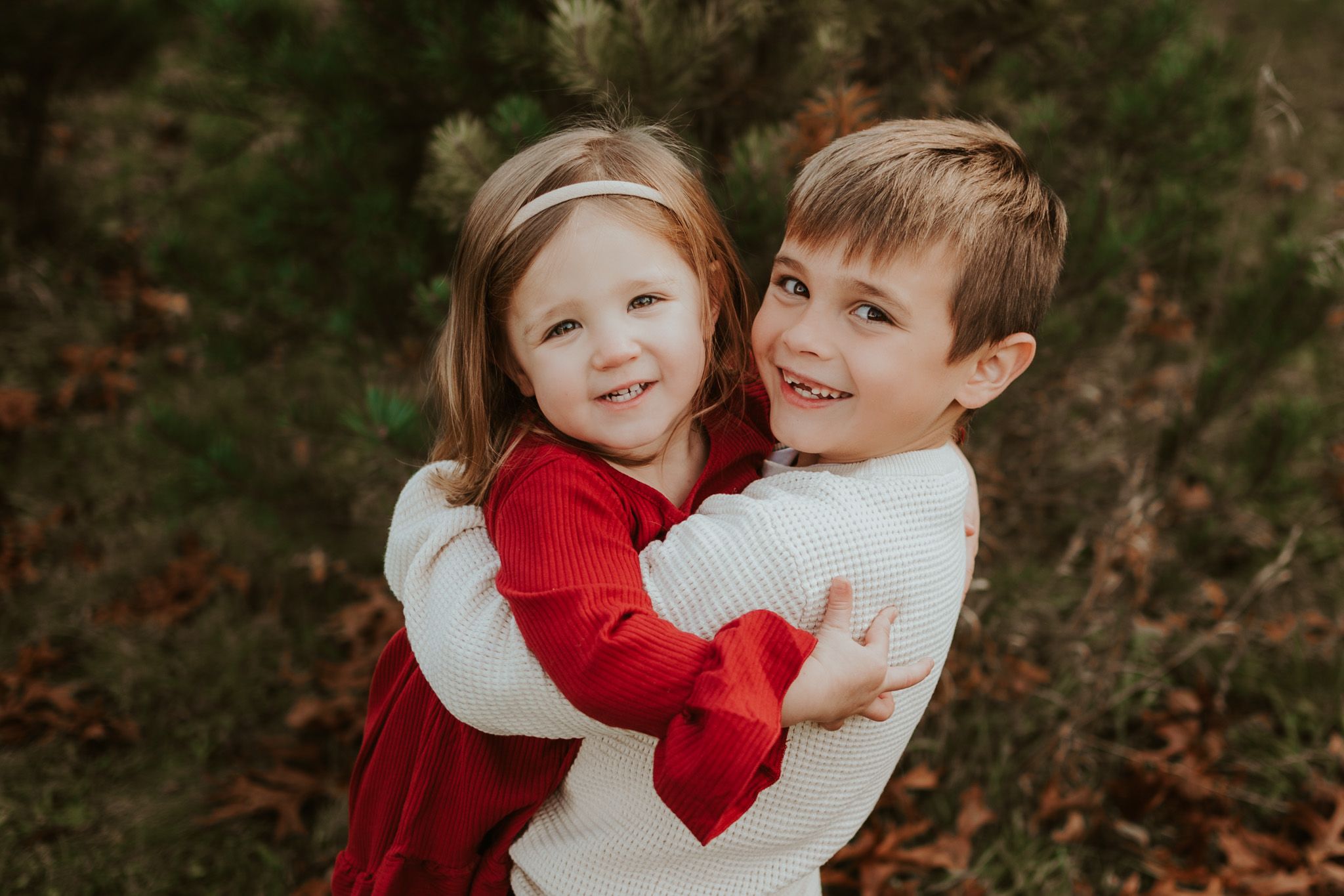 Young girl in red dress and boy in white sweater share a warm hug outdoors among pine trees.