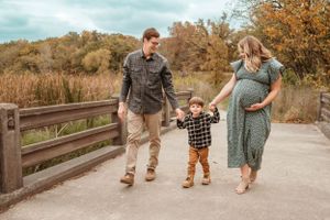 Expecting couple holding hands with toddler son walk on wooden bridge surrounded by autumn foliage outdoors.