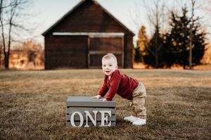 Baby boy in red shirt crawling near a wooden ONE sign outdoors.