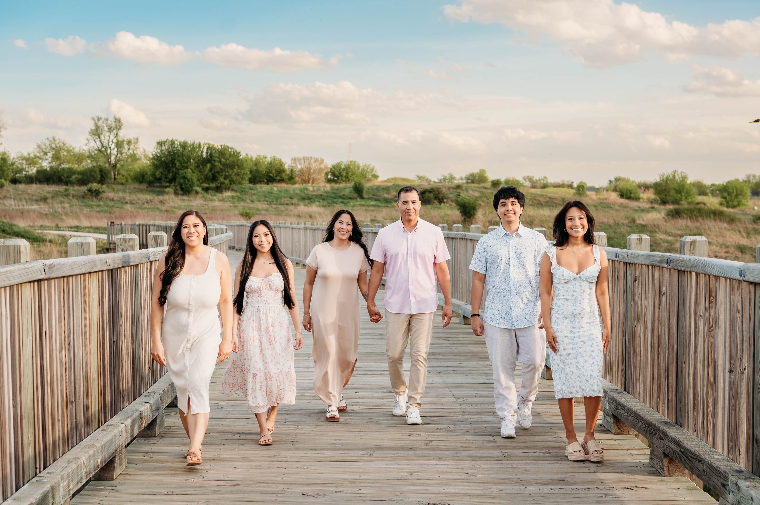 Family of six walking hand-in-hand on a wooden boardwalk near a beach on a sunny day.