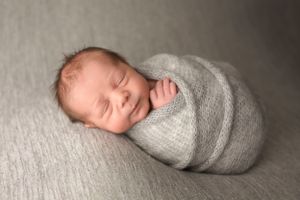 Peaceful sleeping newborn baby swaddled in gray wrap with a gentle smile on soft gray backdrop.