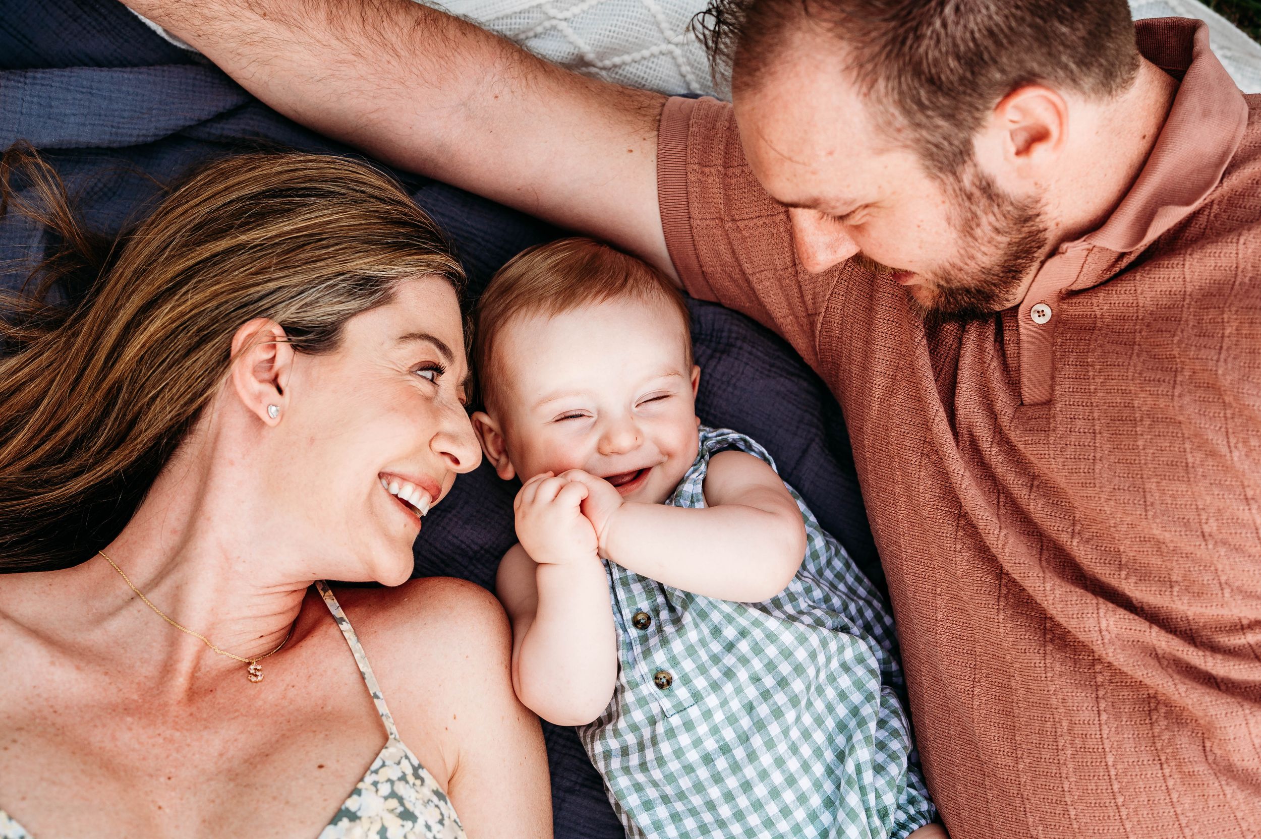 Happy family with smiling baby lying on bed, parents gazing lovingly at laughing infant in gingham outfit.