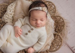 Sleeping newborn baby in cream knit outfit with floral headband in woven basket.
