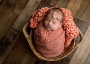 Sleeping newborn baby girl swaddled in pink wrap nestled in a wicker basket with knit blanket.