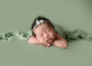 Sleeping newborn baby girl in sage green wrap with floral headband on green backdrop.