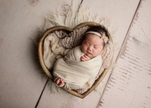 Swaddled newborn baby sleeping peacefully in a wooden heart-shaped bowl with cream wrap.