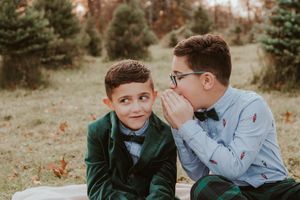 Two boys in formal attire sit on blanket outdoors as one whispers a secret into the other's ear.