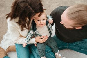 Young parents sitting outdoors on ground smiling at their happy baby boy in plaid shirt and jeans.