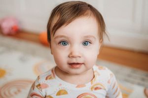 Adorable baby girl with bright blue eyes and brown hair wearing a colorful printed onesie looks at camera.