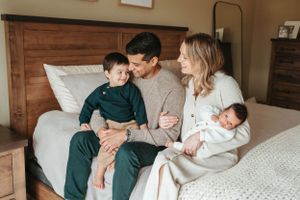 Family of four with newborn baby sitting on a bed, parents smiling at toddler son in cozy bedroom setting.