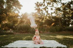 Baby girl in pink dress celebrates first birthday with number one balloon in sunny outdoor park setting.