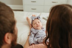 New parents lovingly gaze at their sleeping newborn baby wearing a grey bow headband on a white bed.