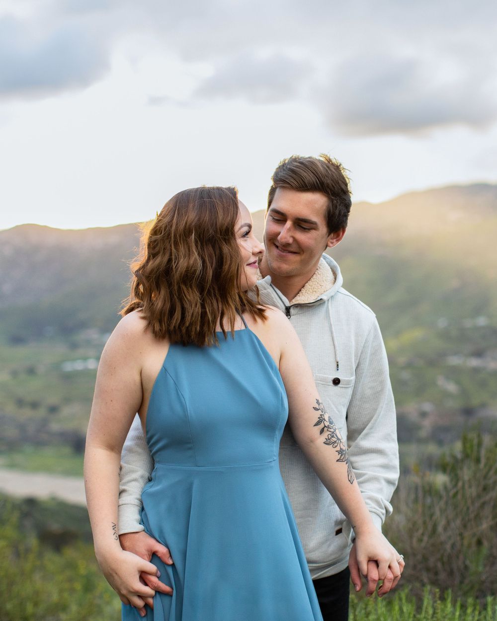Couple in blue dress and grey hoodie sharing an intimate moment with mountains behind them.