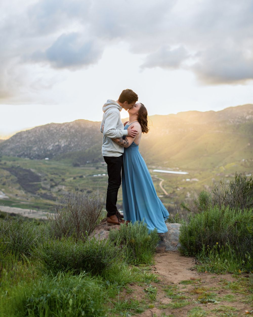 Couple kissing on hilltop trail during golden hour with scenic mountain backdrop.