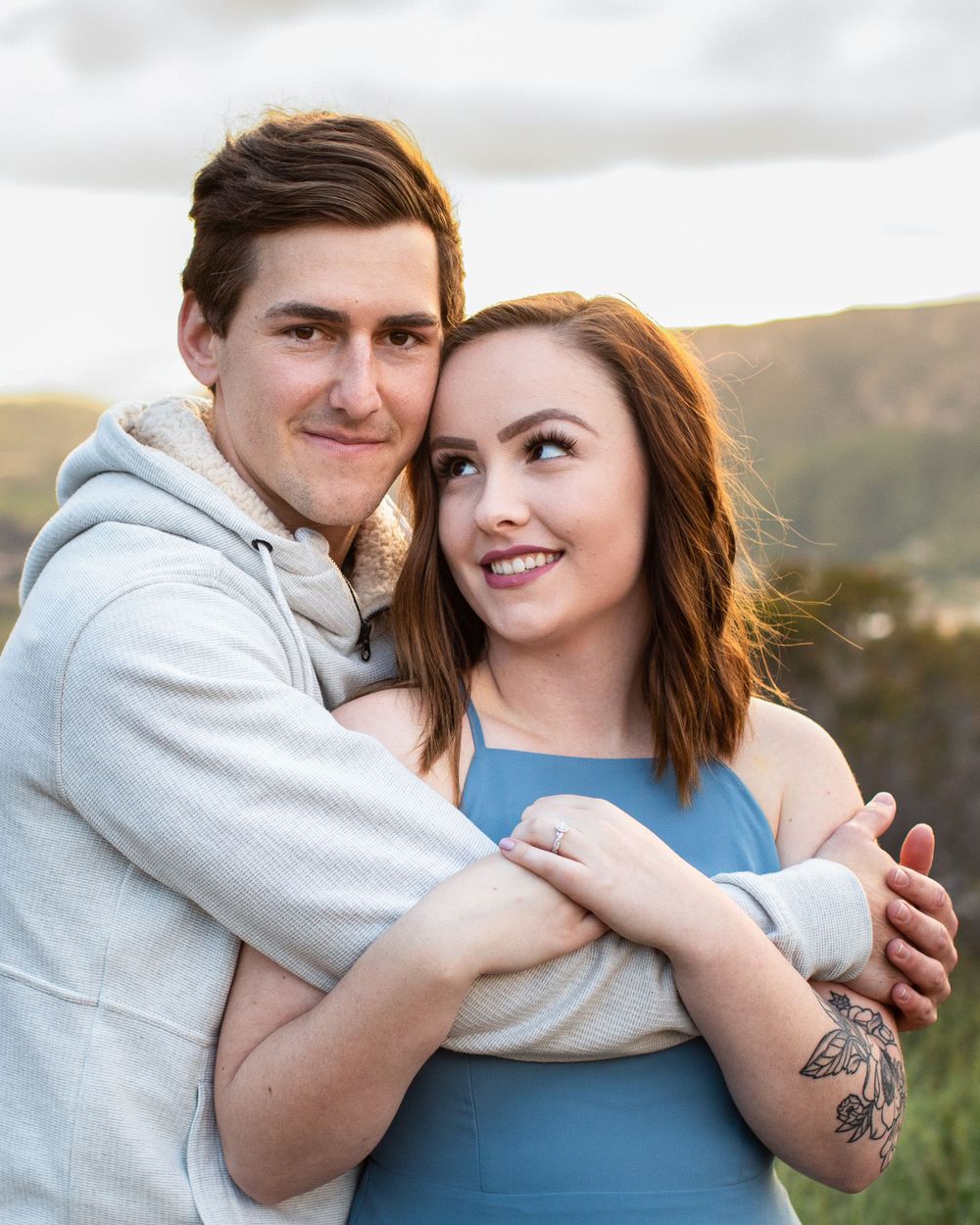 Couple in casual attire smiling and embracing with scenic mountain view behind them.