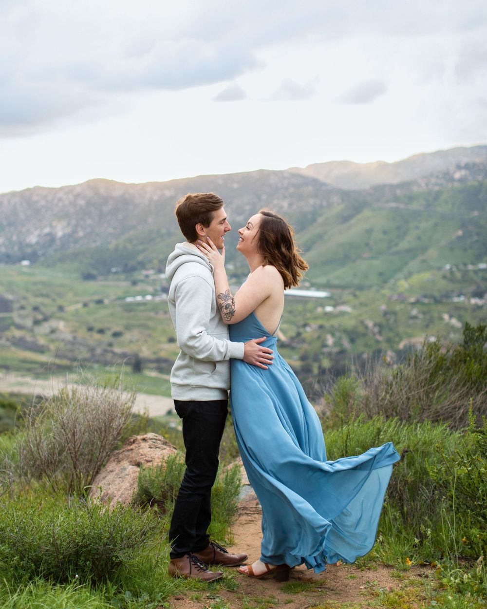 Couple in flowing blue dress and grey sweater embracing on mountain path.