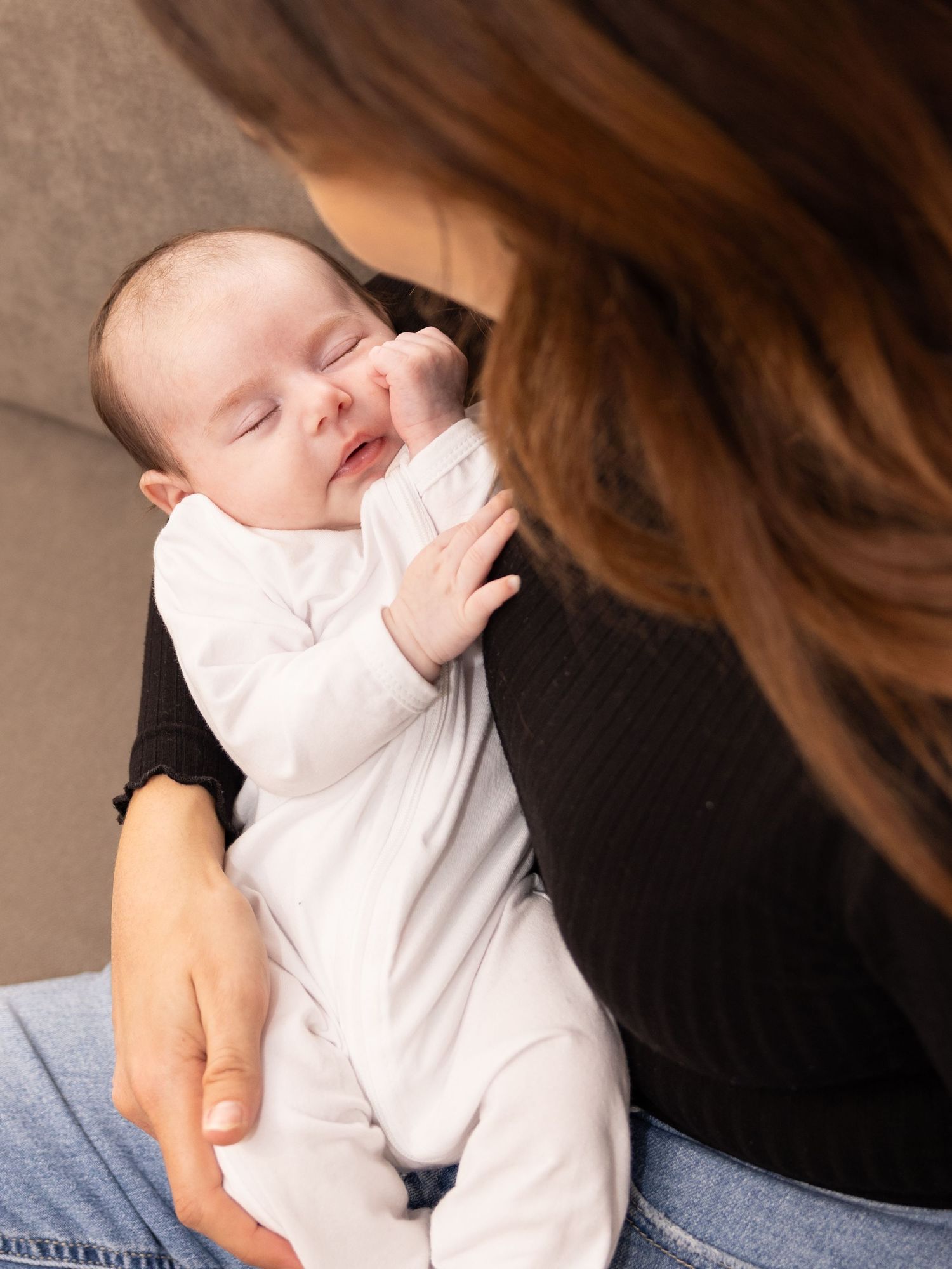 Baby in white onesie reaching up while being held on couch.