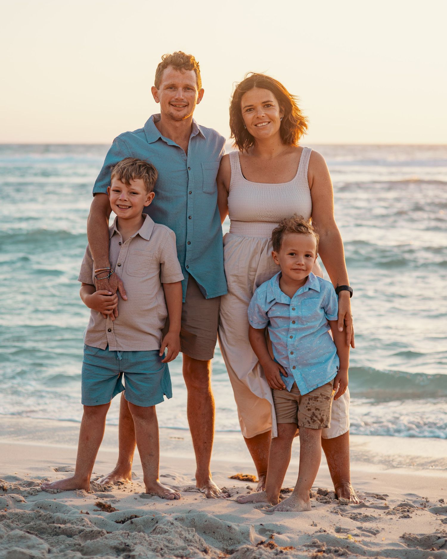 A family in coordinated blue and beige outfits poses together at sunset on a beach with ocean waves in the background.