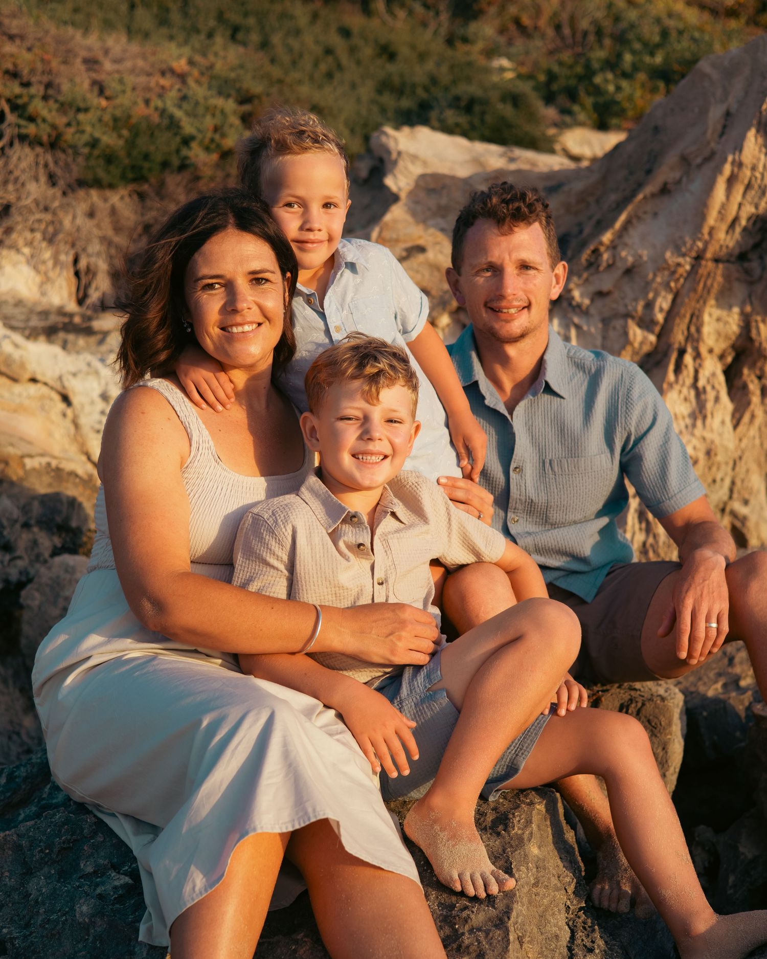 A family of four poses together on coastal rocks during golden hour sunset lighting.