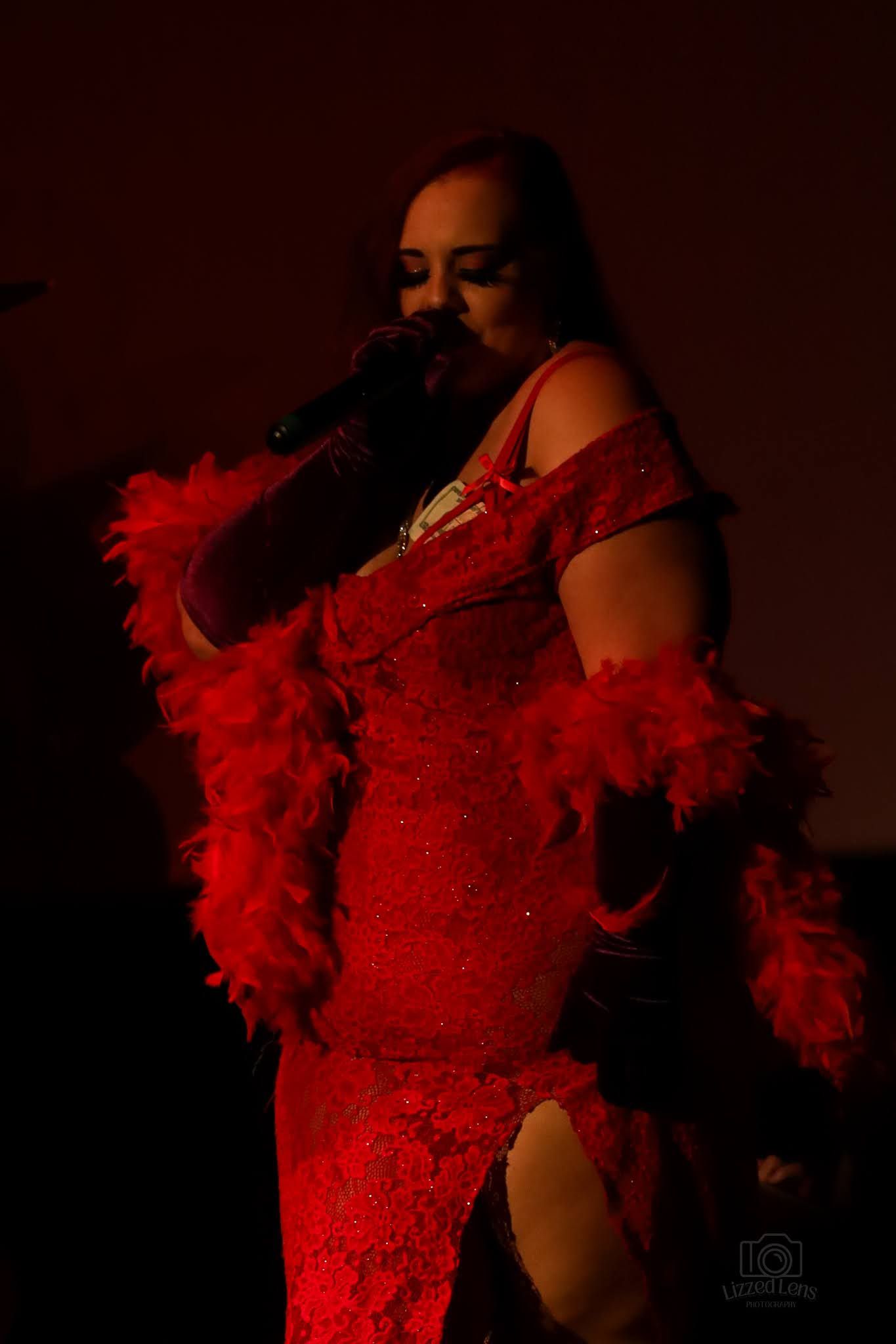 A performer in a sparkling red gown with feather boa sings on a dimly lit stage.