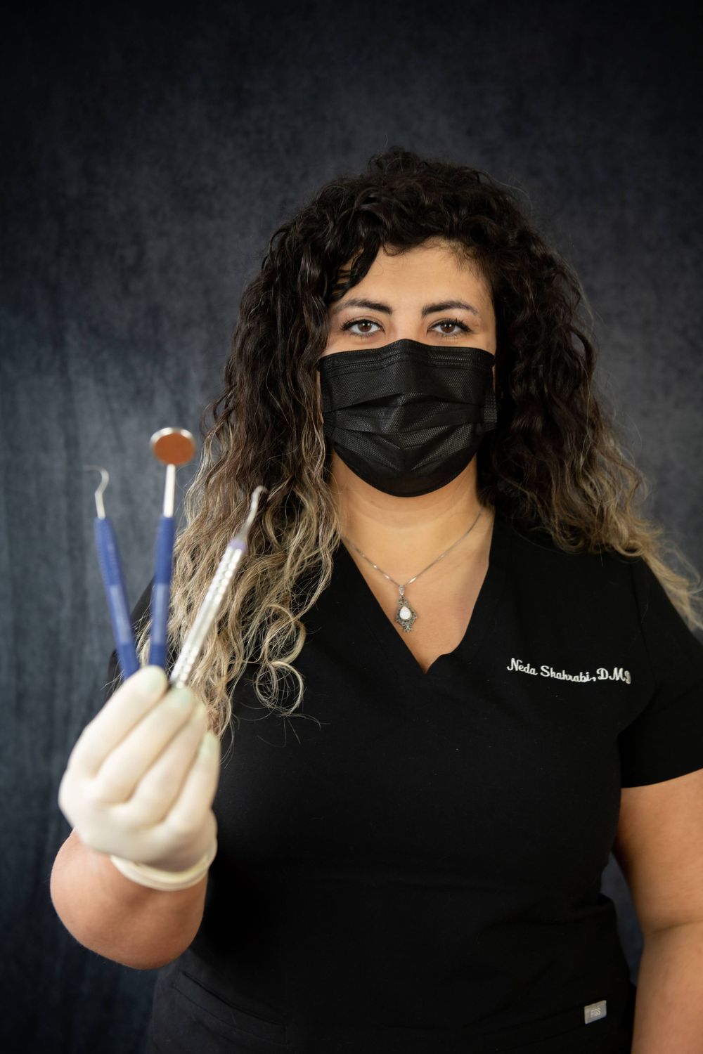 A healthcare professional in a black mask and gloves holds dental tools against a dark background.