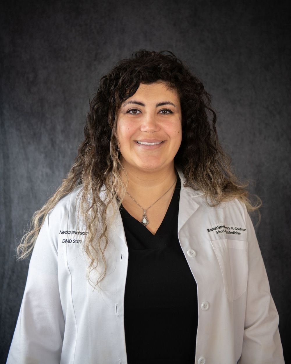 Medical professional wearing a white coat and black top poses against dark studio backdrop for professional headshot.