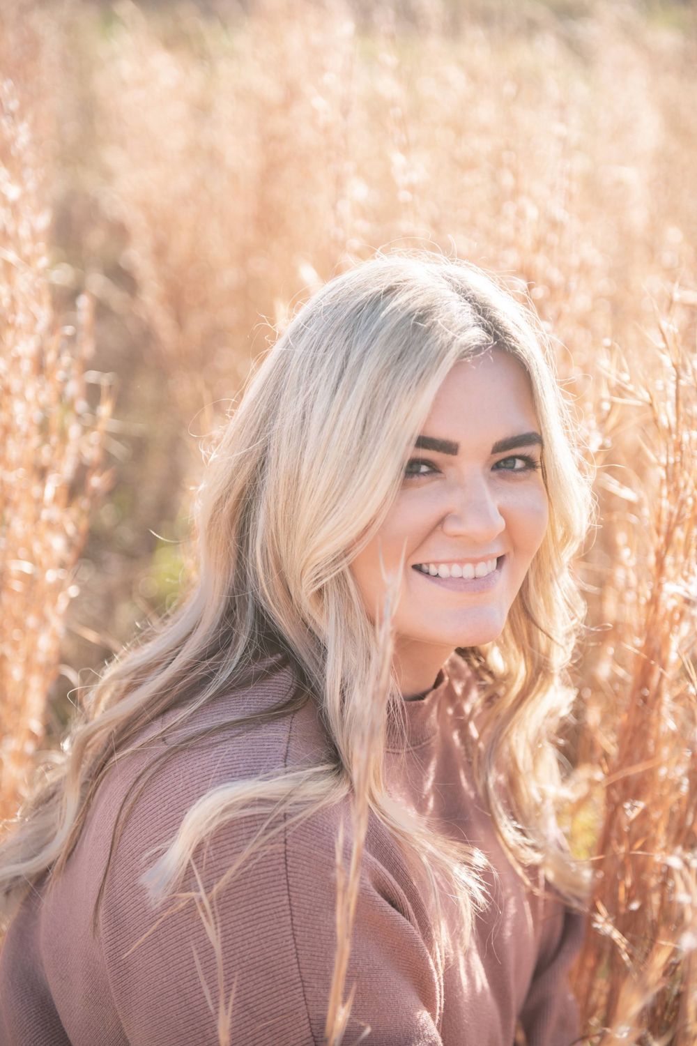 Smiling person with long blonde hair poses in a brown sweater against sunlit autumn grasses during golden hour.