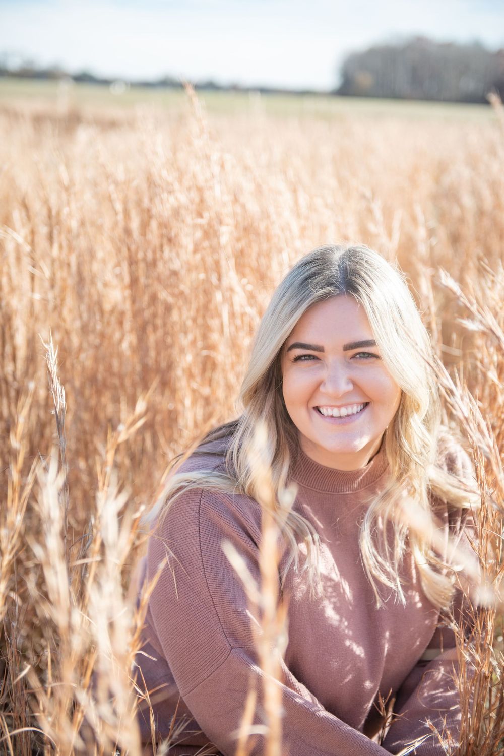 Smiling woman wearing mauve sweater sitting in golden wheat field on sunny autumn day.