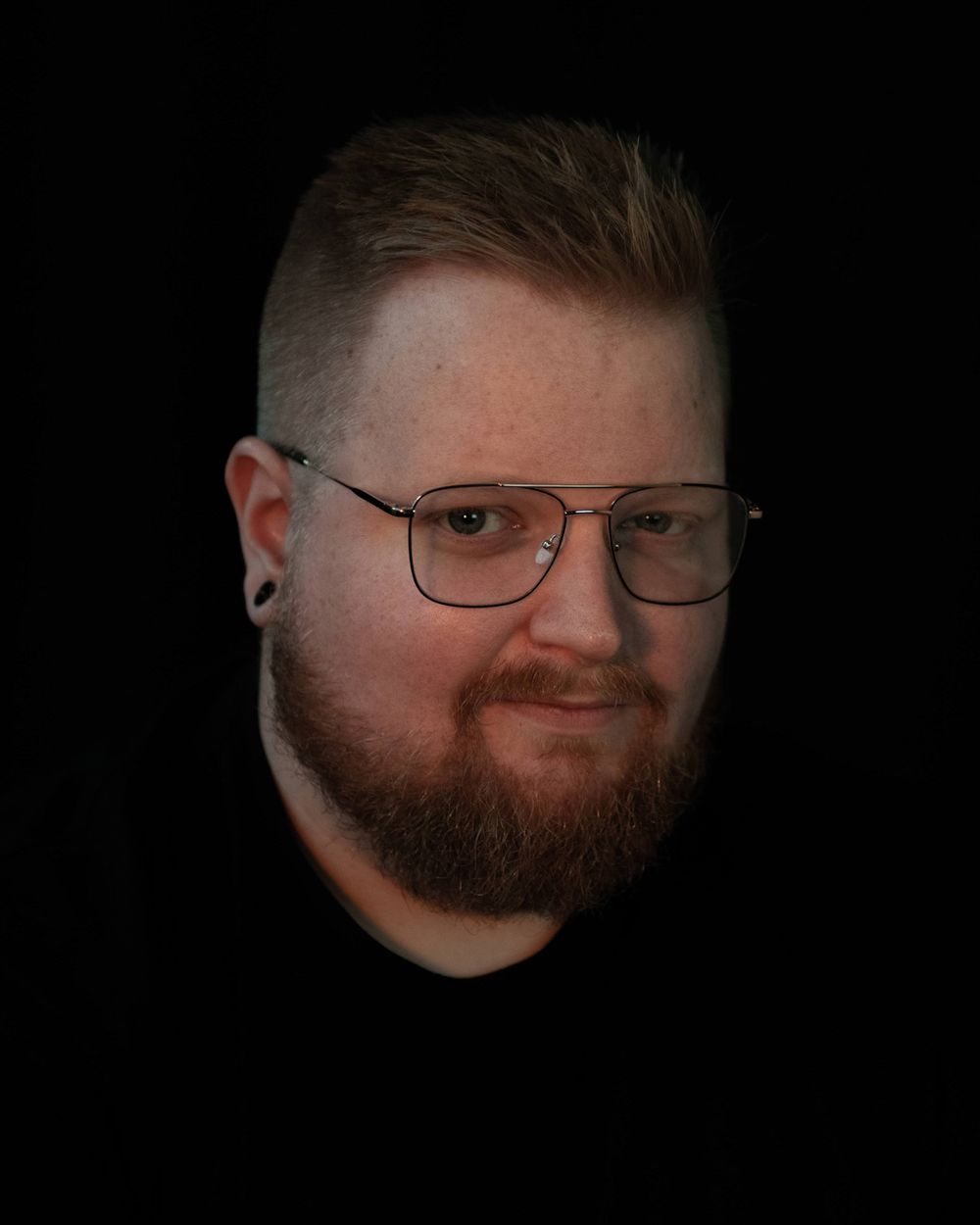 Close-up headshot with glasses against dark background.