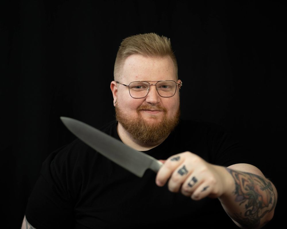 Professional portrait of a person with glasses and beard holding a chef's knife against dark background.