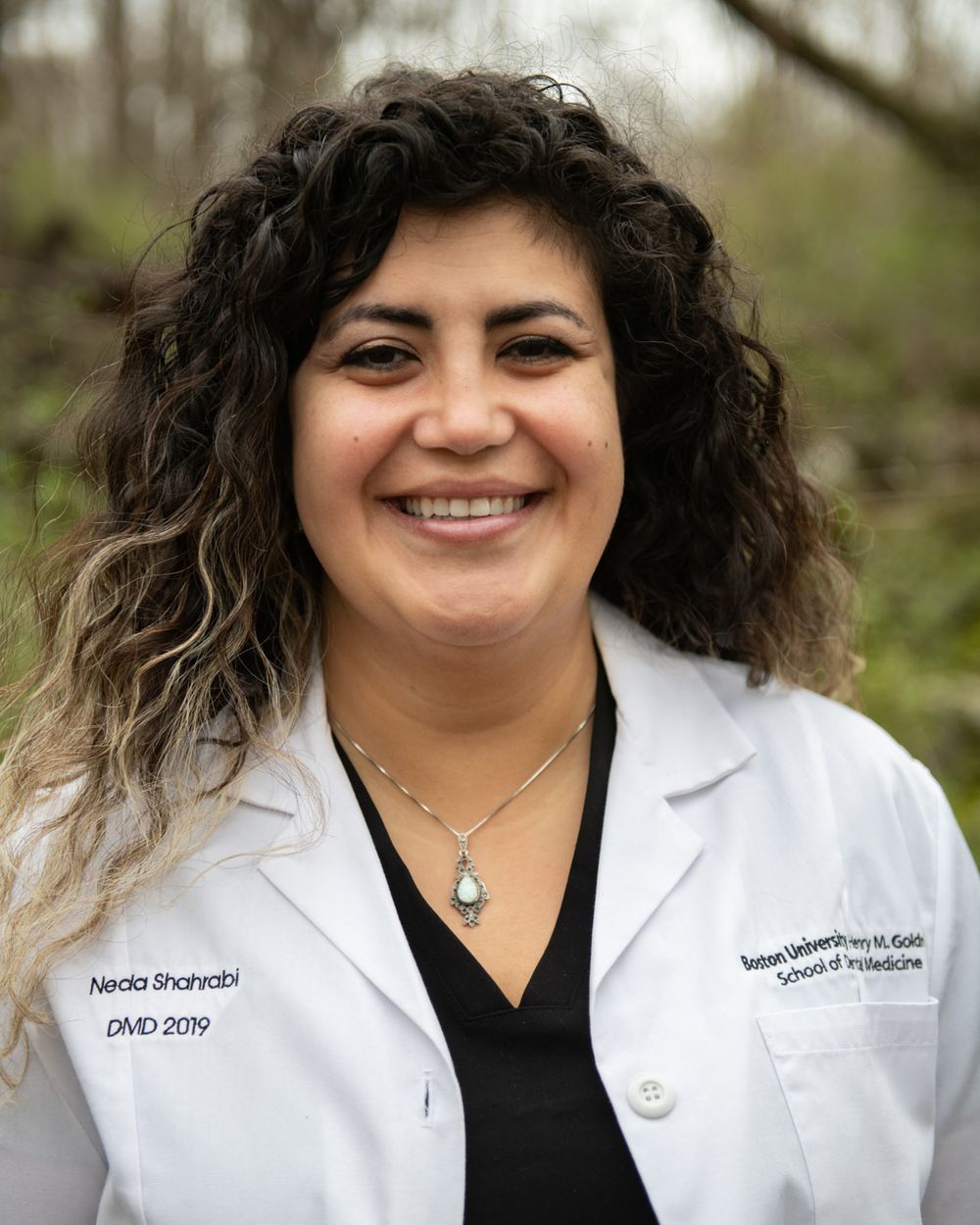 Professional portrait of person in white medical coat and black top with long curly hair outdoors.