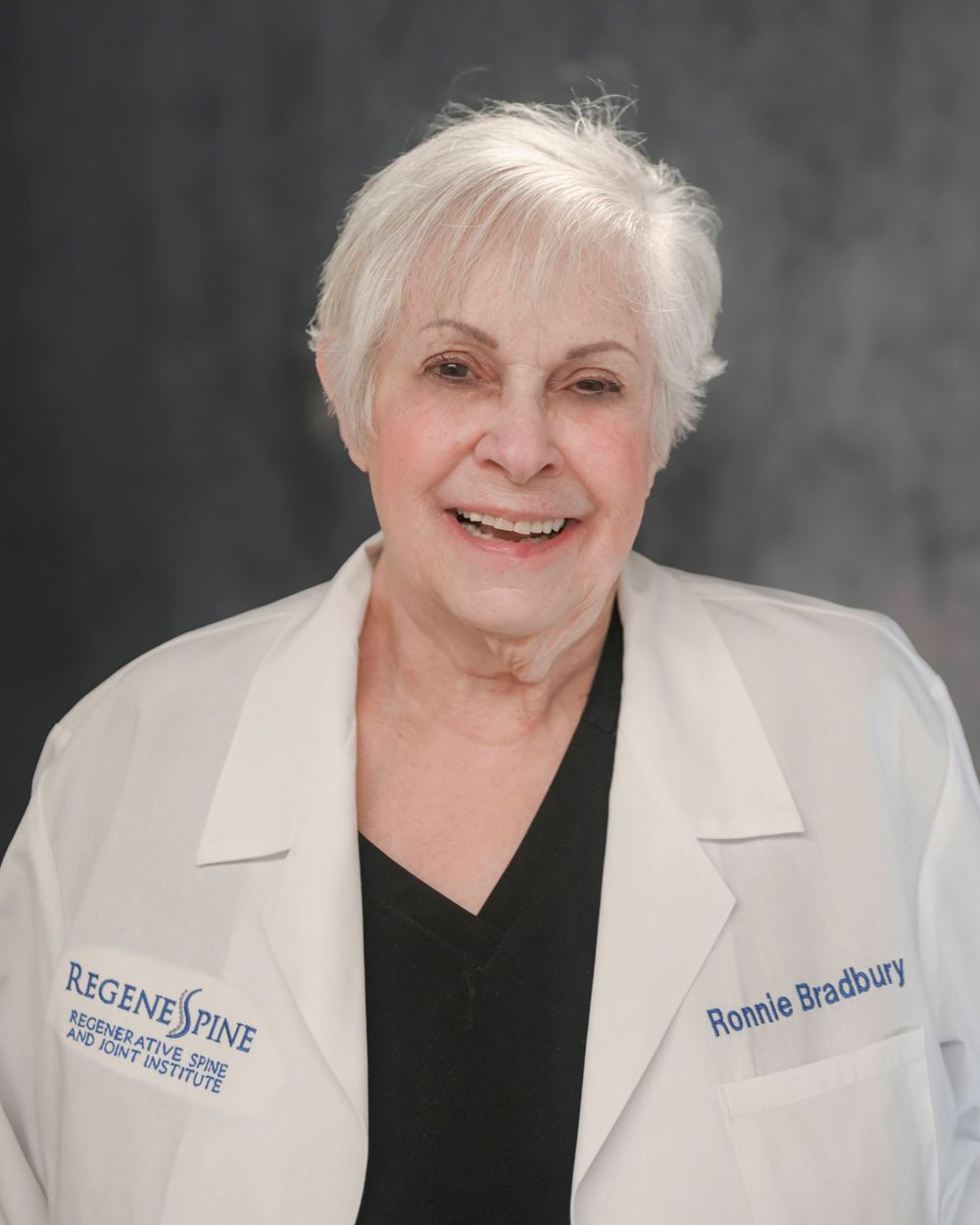Professional headshot of person in white blazer and black top against grey background.