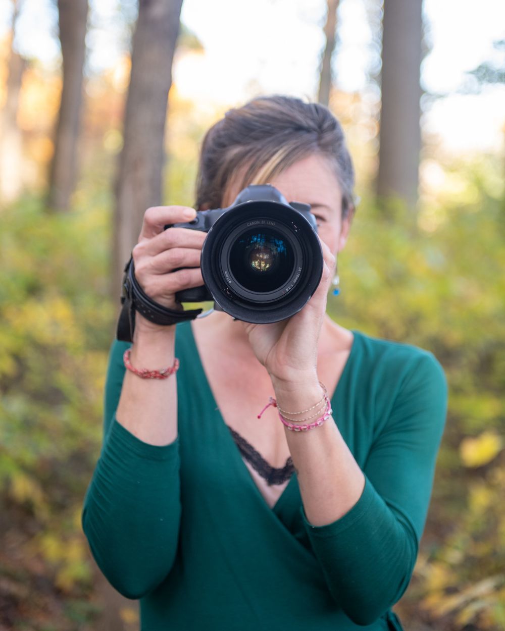 Photographer wearing green sweater aims professional DSLR camera in autumn forest setting.