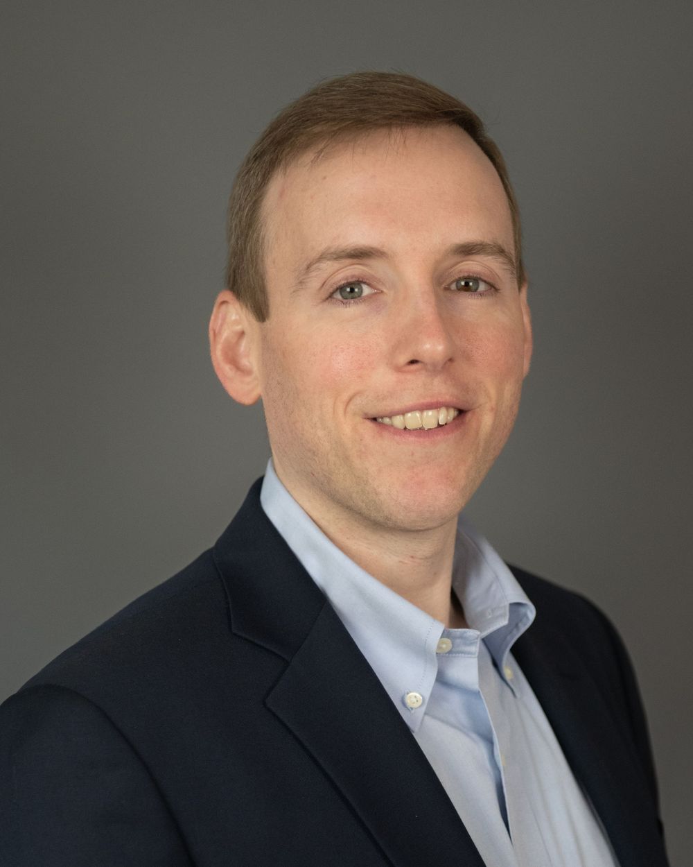 Professional headshot of person in navy blazer and light blue button-down shirt against gray background.