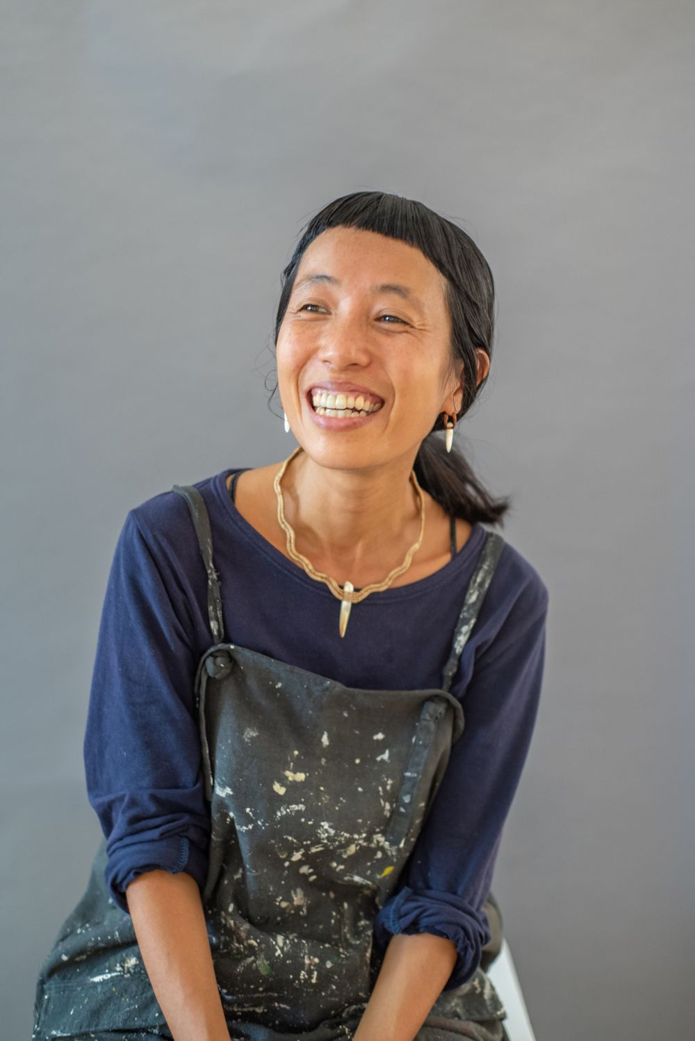 An artist wearing paint-splattered overalls and a navy blue shirt smiles warmly against a gray background.