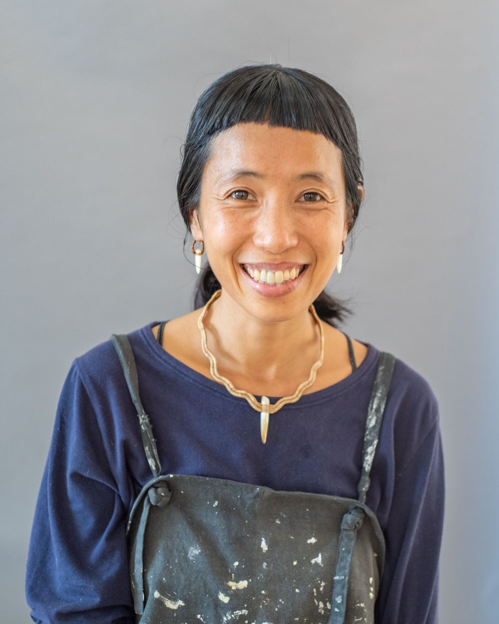 A cheerful portrait of someone in a navy blue top and artist apron against a gray backdrop.