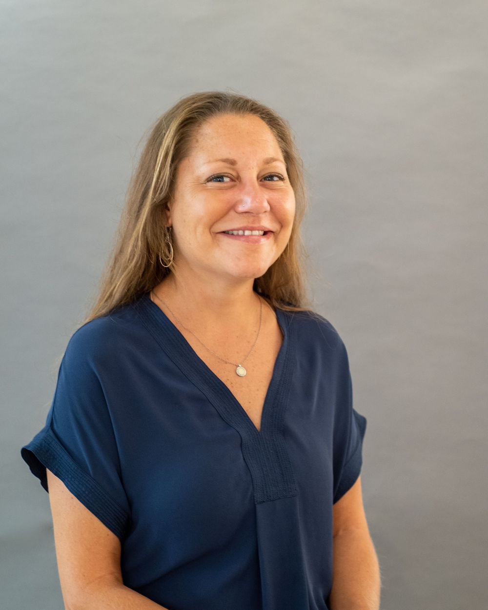 Professional headshot of someone wearing a navy blue v-neck blouse against a light gray background.