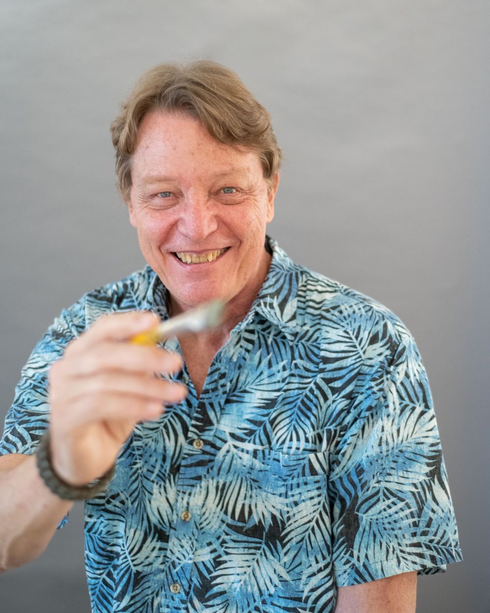Someone wearing a blue tropical print Hawaiian shirt holds a cocktail glass while smiling against a gray background.