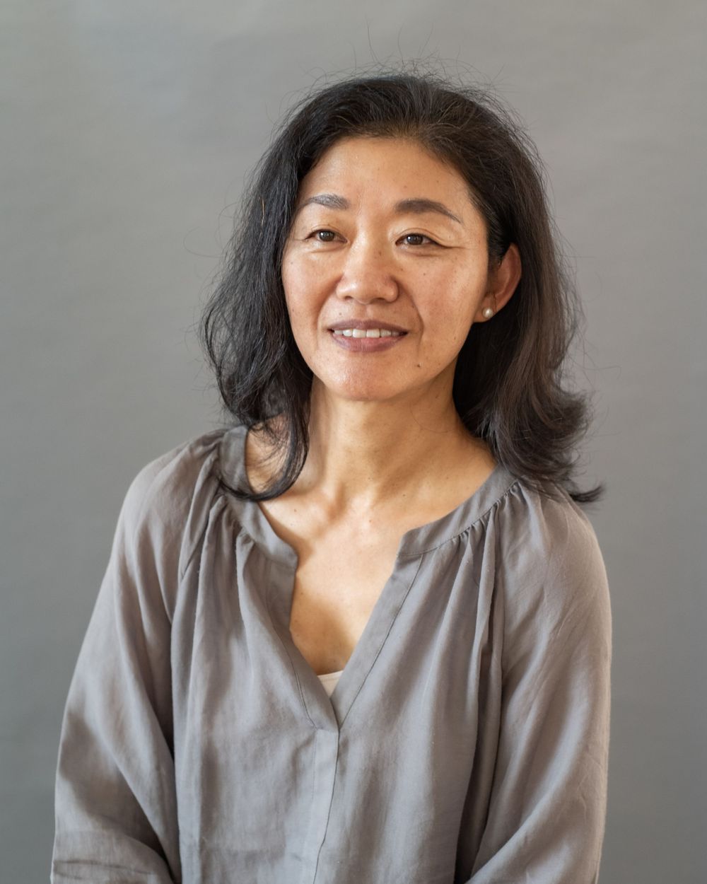 A person with dark hair wearing a soft gray top smiles warmly against a neutral background.