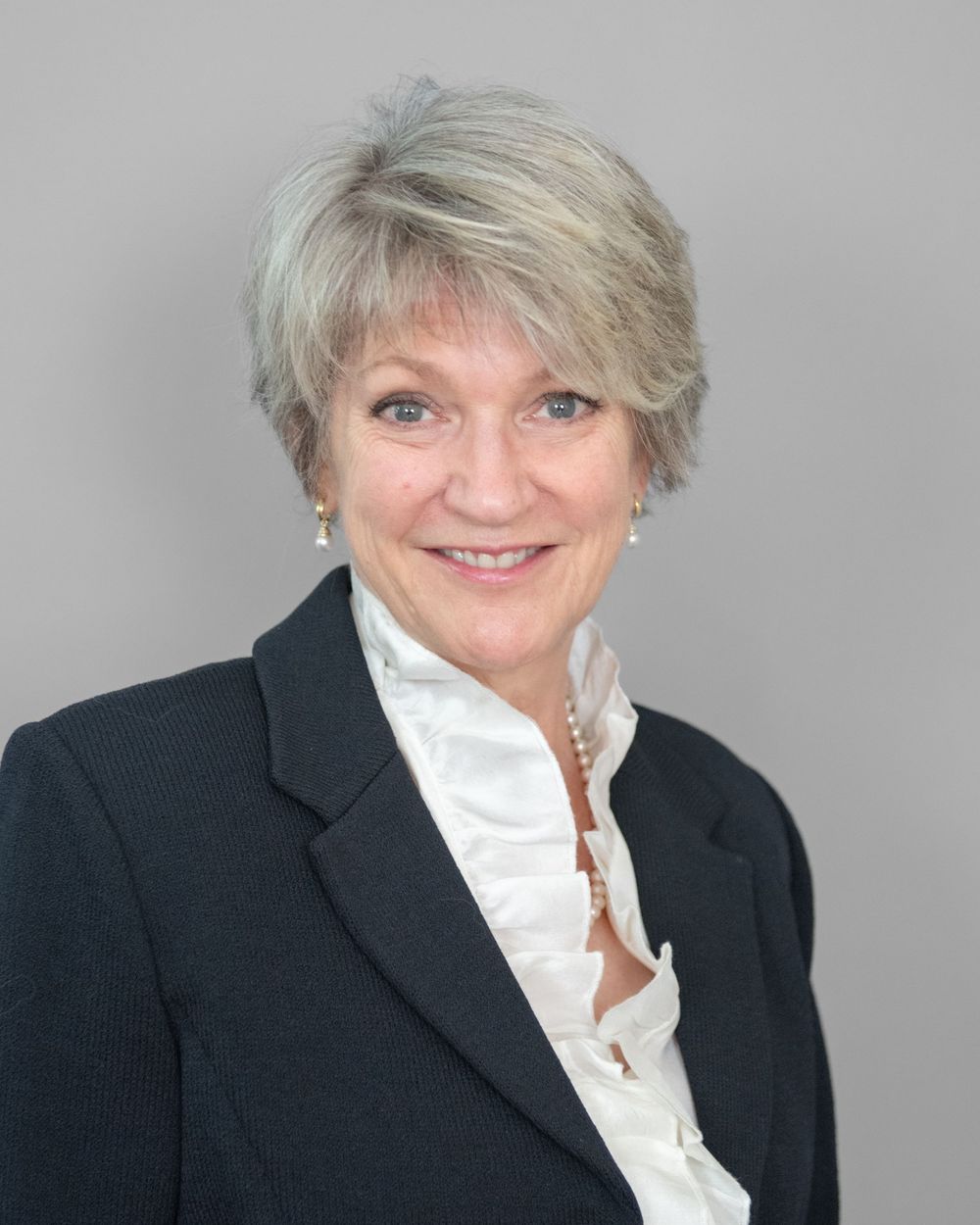Professional business headshot showing a person in a black blazer and white blouse with silver-gray hair against a gray background.