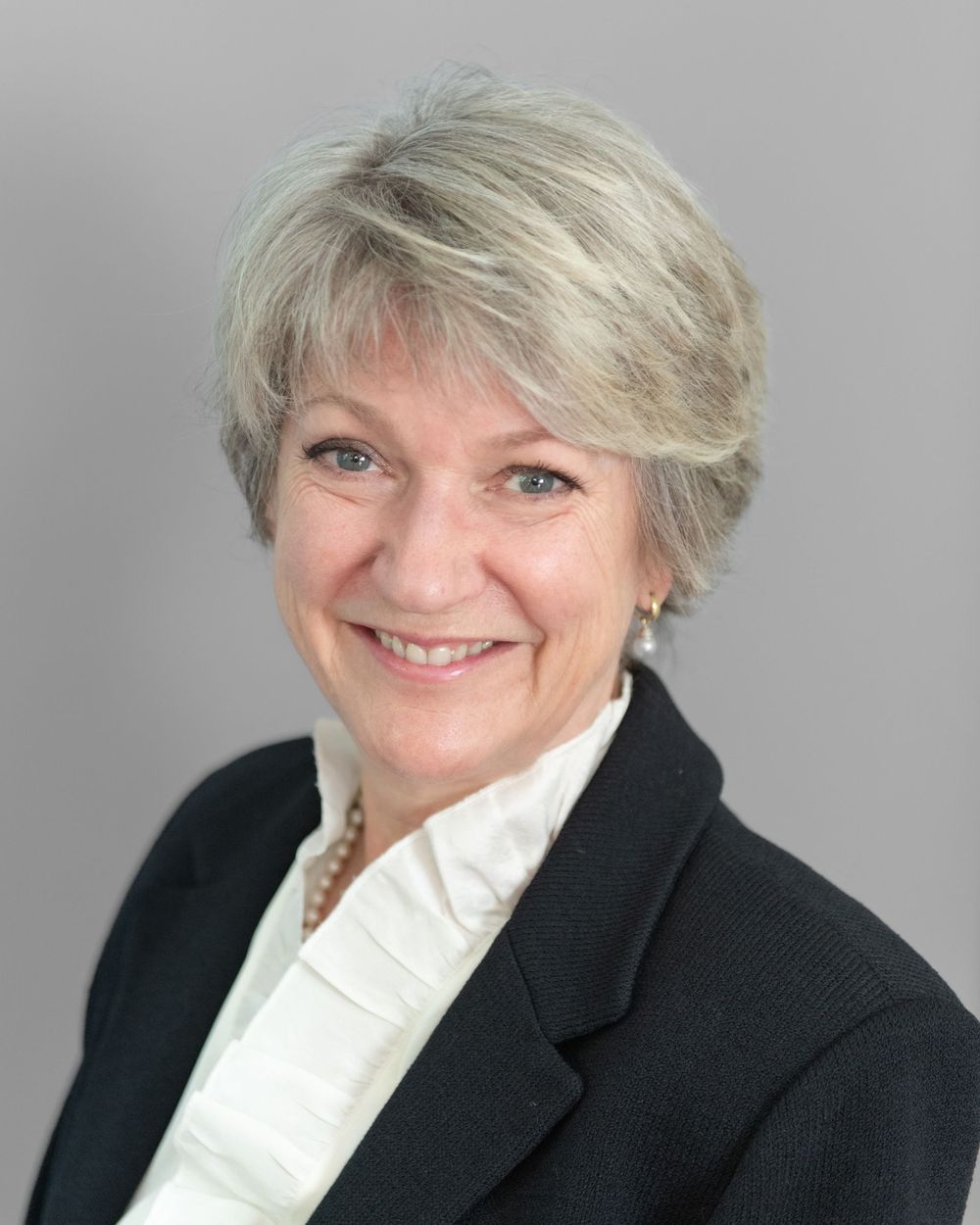 Professional headshot of a person wearing a black blazer and white collared shirt against a gray background.