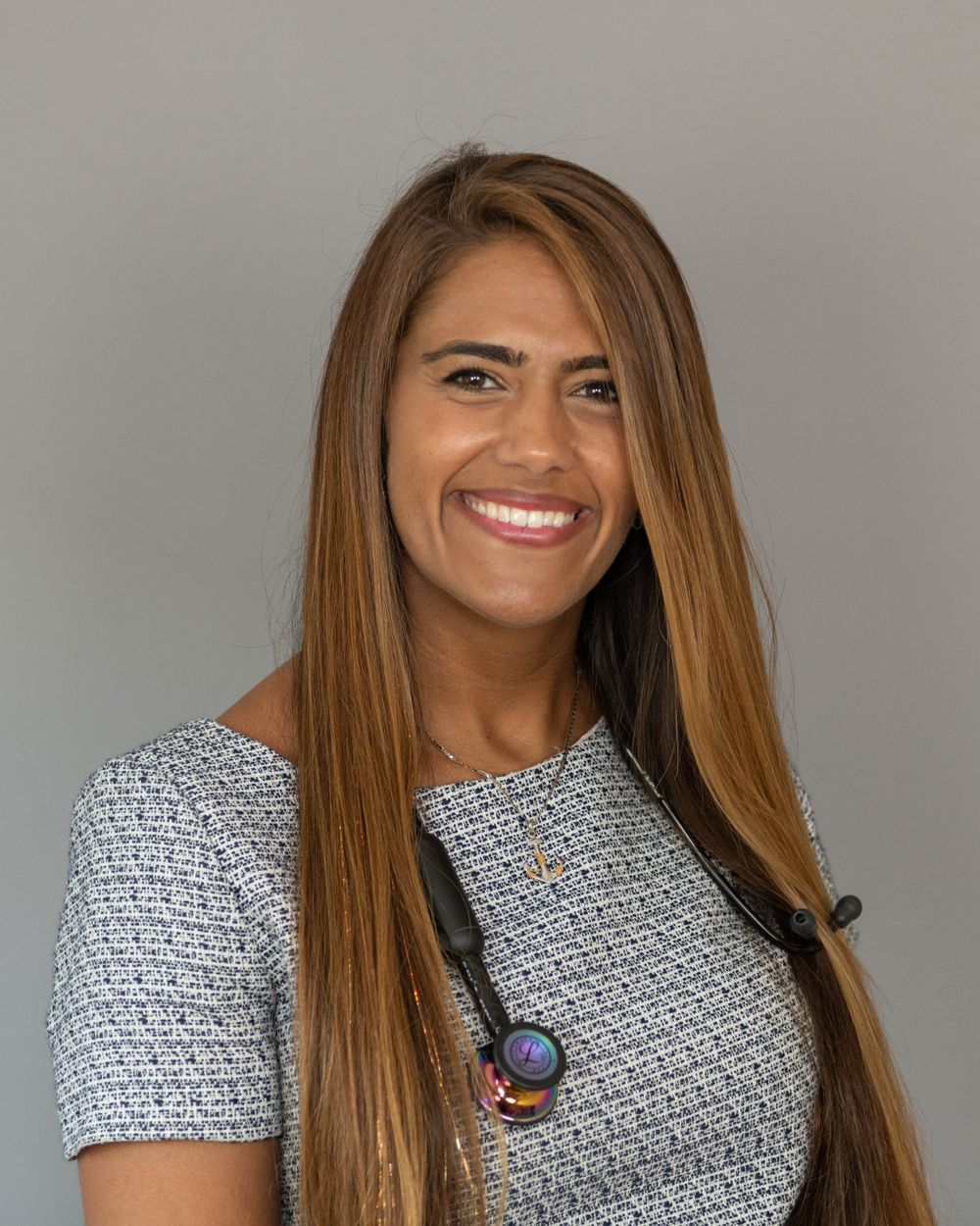 Professional headshot of a smiling person with long brown hair wearing a gray patterned top and pendant necklace.