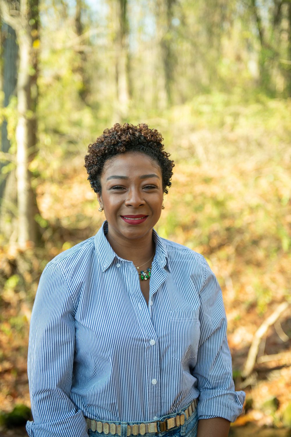 A person wearing a blue striped button-down shirt smiles while standing in a sunlit autumn forest.