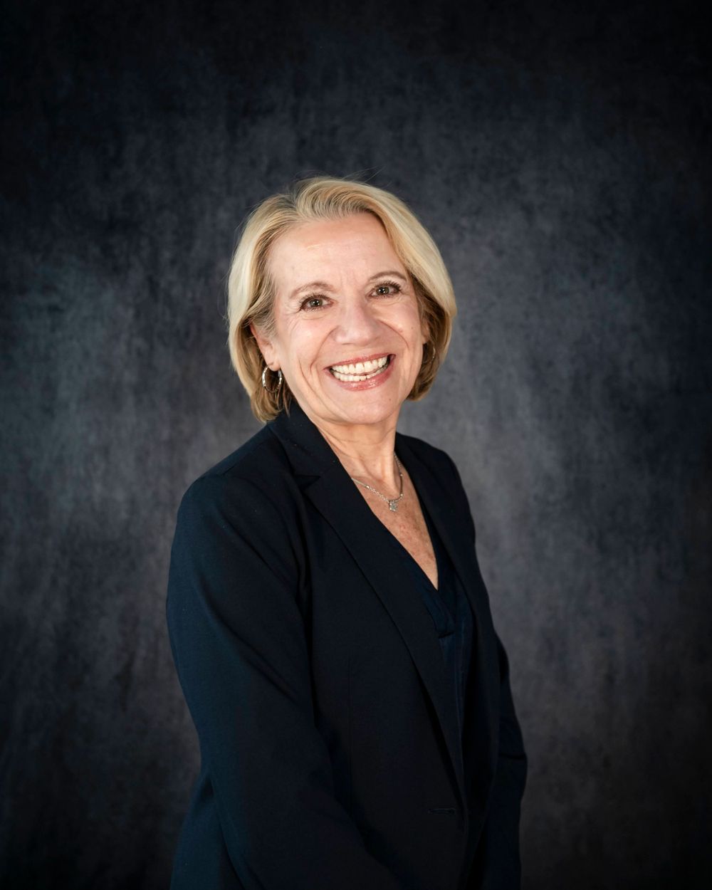 Professional studio portrait of a person in a black blazer smiling warmly against a dark textured background.