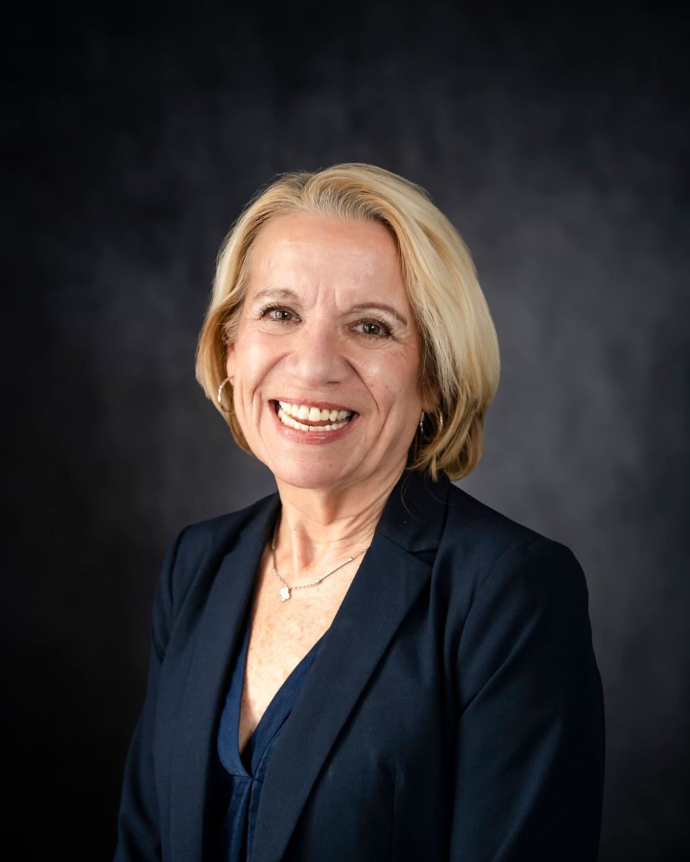 Professional portrait of a person in a navy suit and necklace against a dark backdrop.