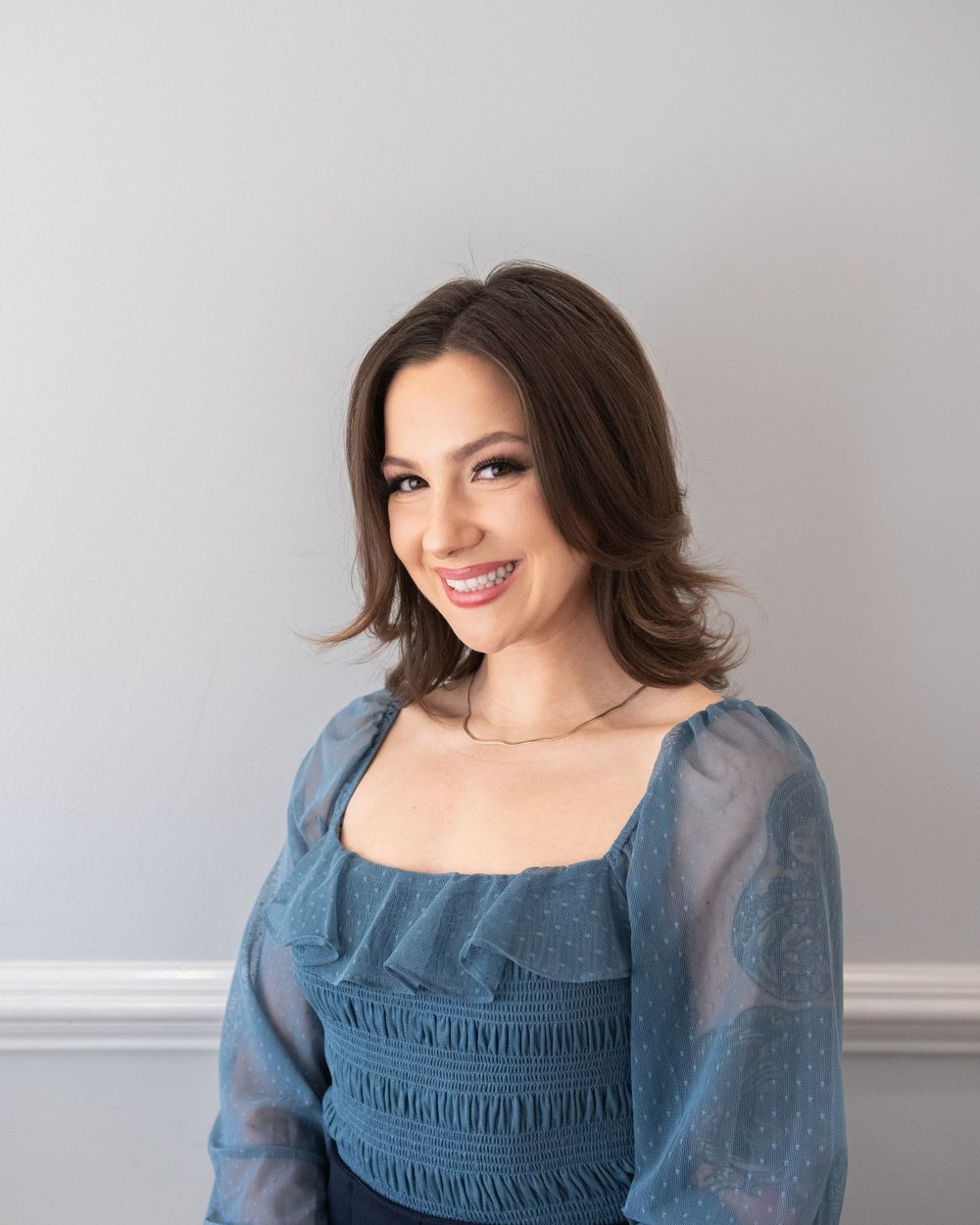 Cheerful woman wearing a blue ruched bodice dress with sheer puff sleeves poses against a gray wall.