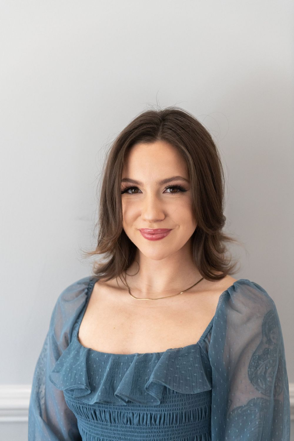 A portrait of someone wearing a blue lace blouse with puff sleeves smiles warmly against a light gray background.