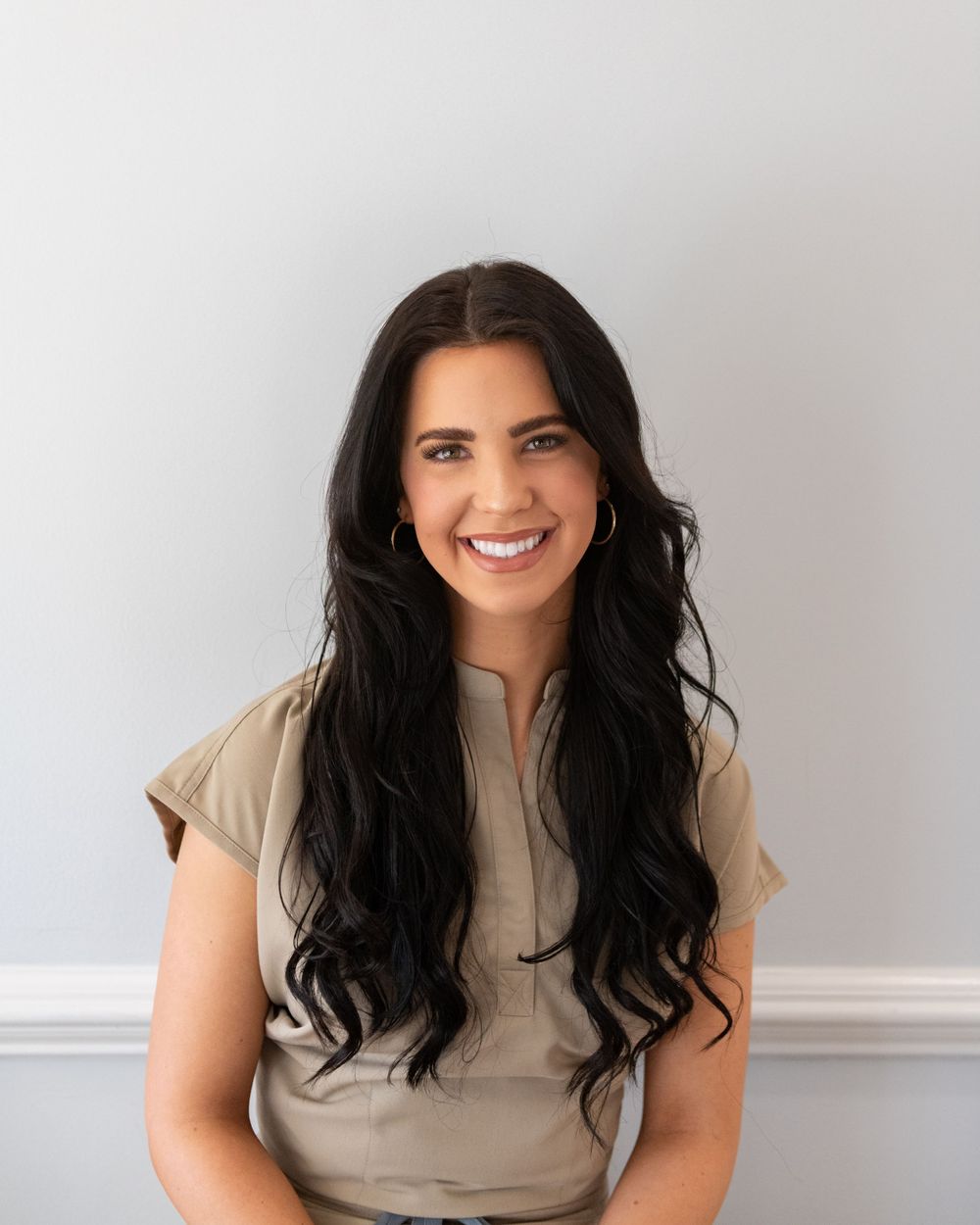 Professional portrait of a person with long dark wavy hair wearing a beige top against a light background, smiling warmly.