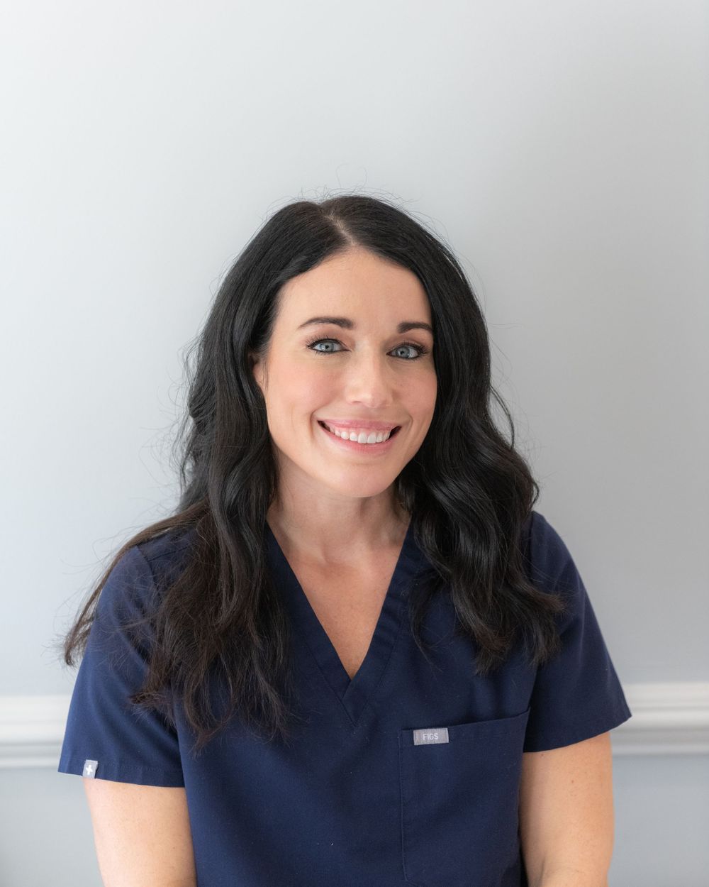Healthcare worker in navy blue scrubs with long dark hair against a light background.